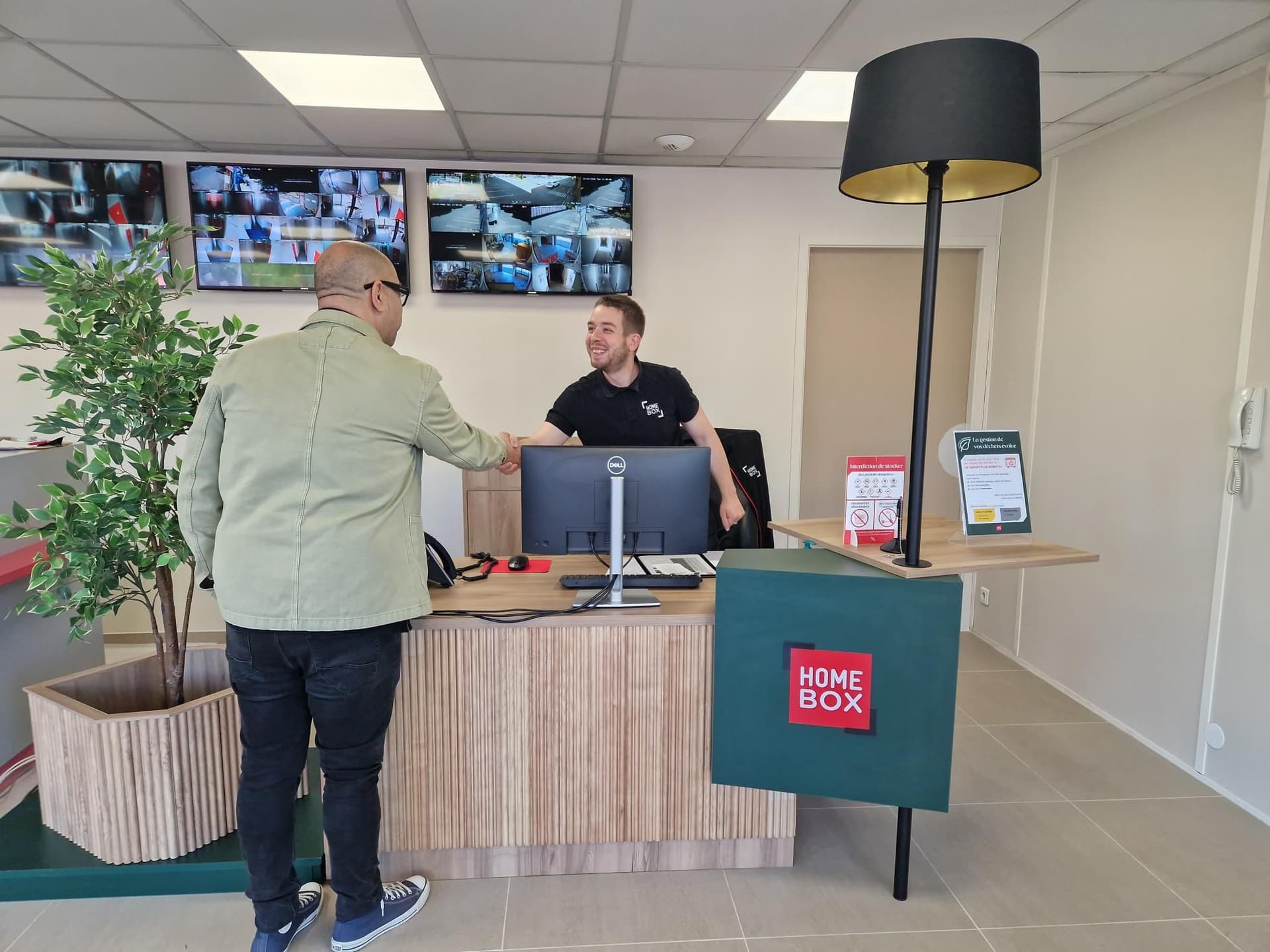 Accueil avec comptoir, écrans de vidéosurveillance et conseiller saluant un client du centre HOMEBOX Evry-Courcouronnes
