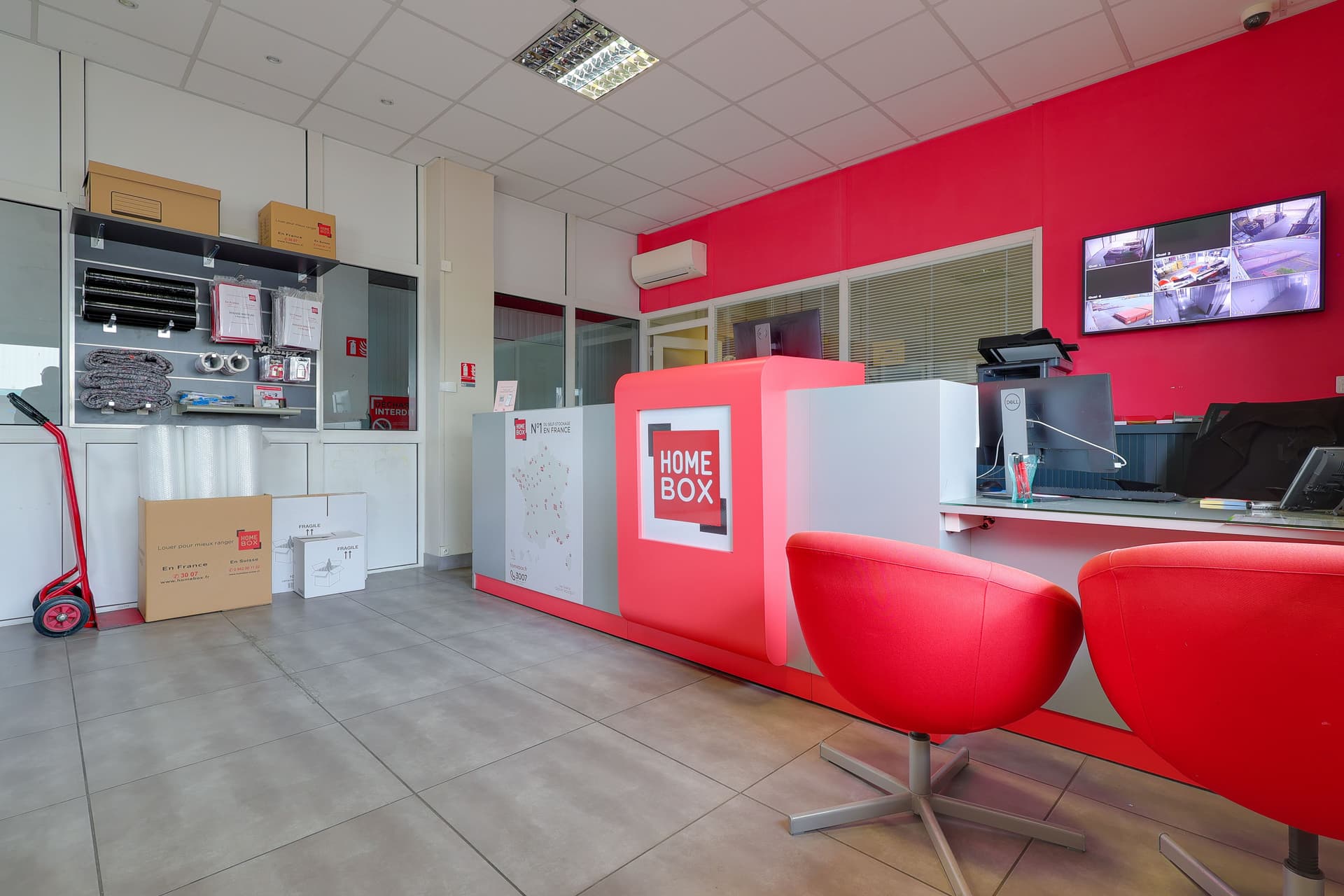 Espace d'accueil avec banque rouge HOMEBOX, fauteuils, écrans de vidéosurveillance et cartons de déménagement du centre HOMEBOX Toulouse Fondeyre