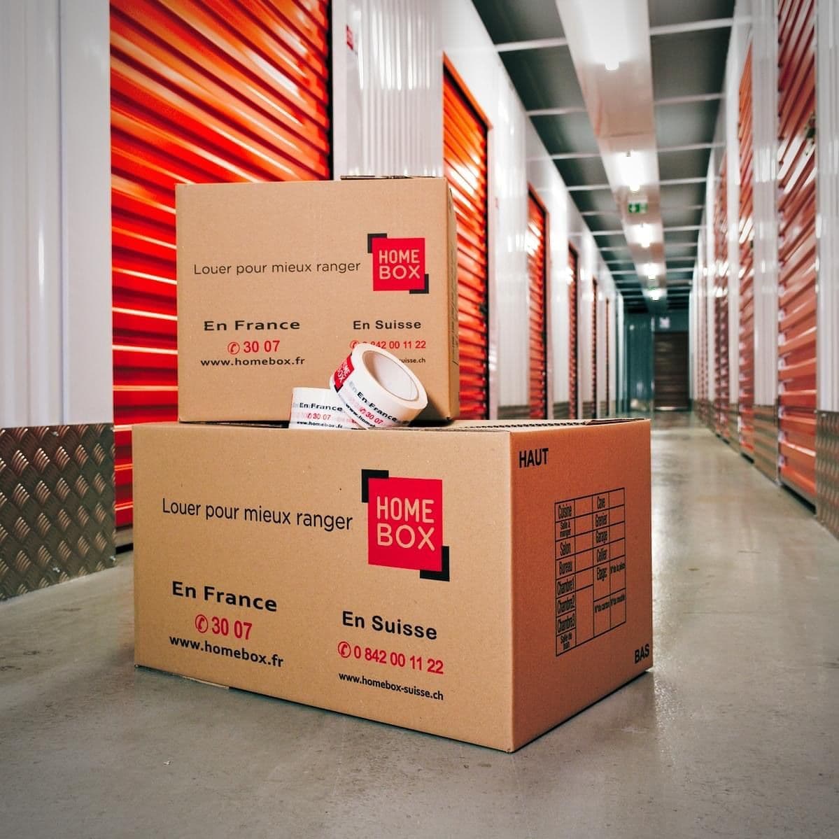 Cartons de déménagement HOMEBOX et rouleau d'adhésif posés dans un couloir de boxes rouges du centre HOMEBOX Niort