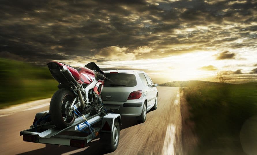 Moto rouge arrimée sur une remorque tractée par une voiture au coucher du soleil