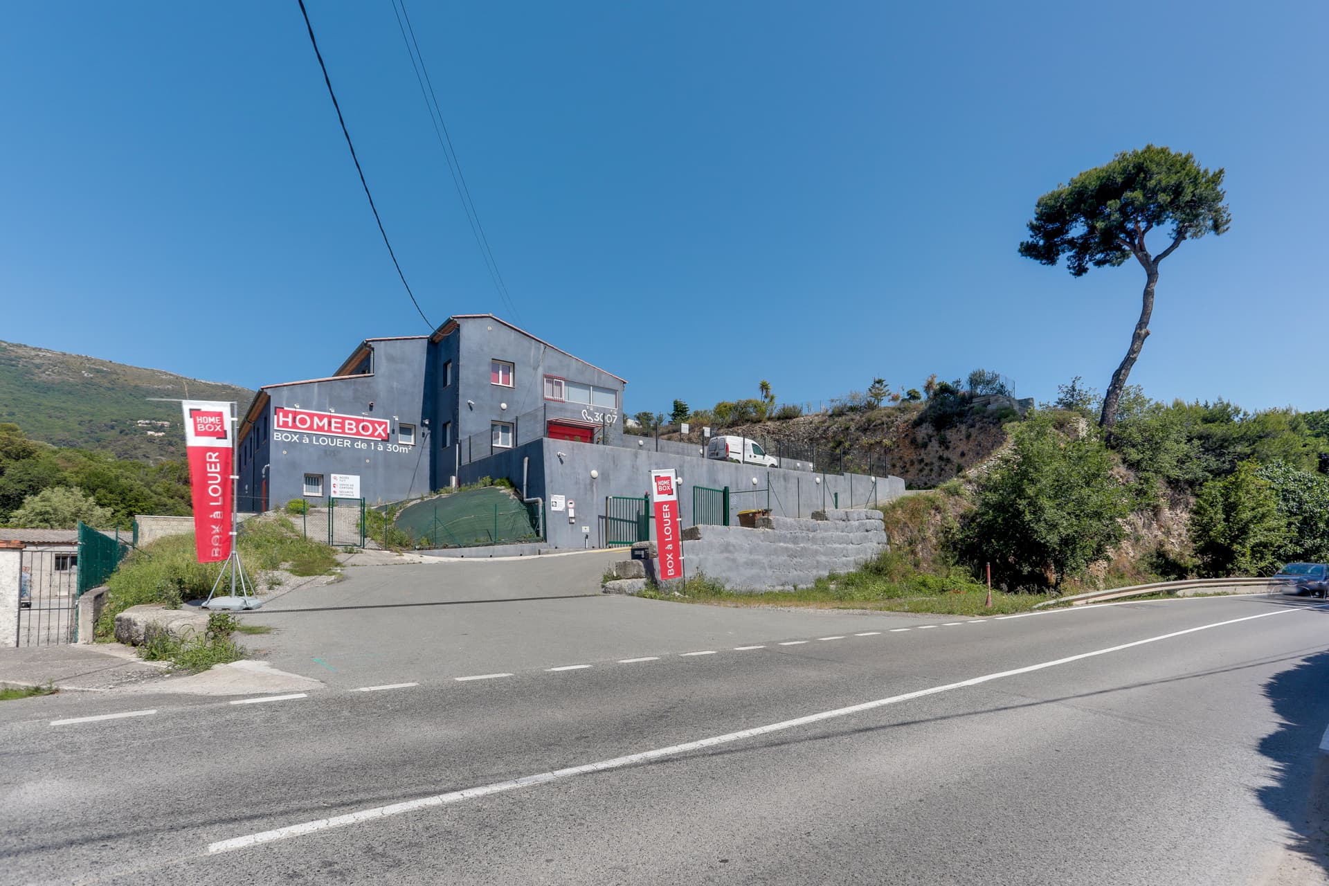 Bâtiment gris en surplomb avec enseigne HOMEBOX et kakémonos en bord de route, HOMEBOX Nice-Carros, 650 route de la Baronne à Gattières
