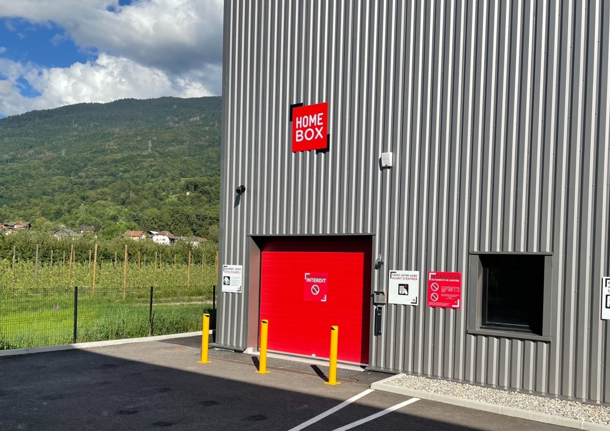 Porte d’accès rouge du centre HOMEBOX Albertville avec poteaux de protection jaunes.