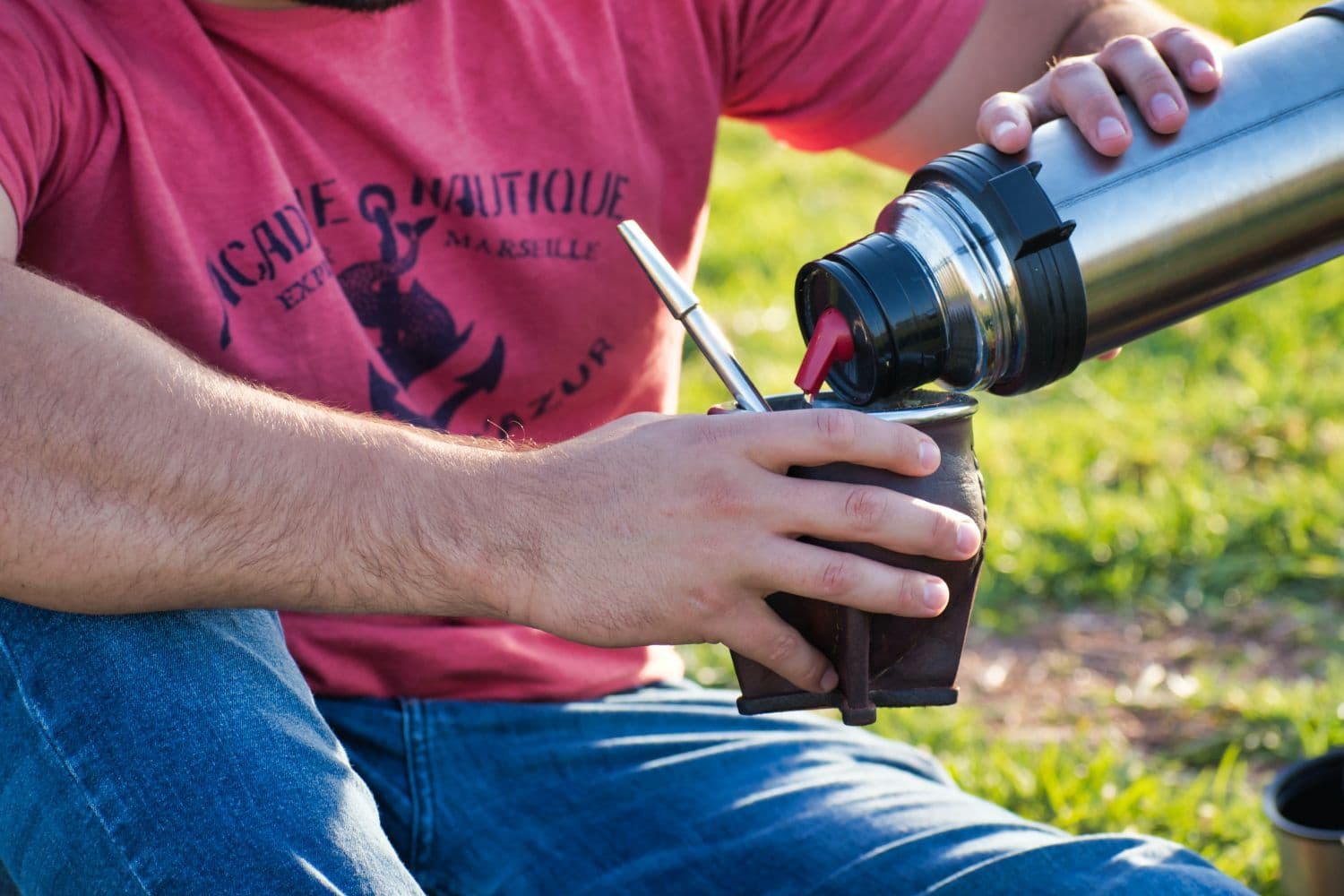 Personne en t-shirt et jean se servant une boisson grâce à un thermos