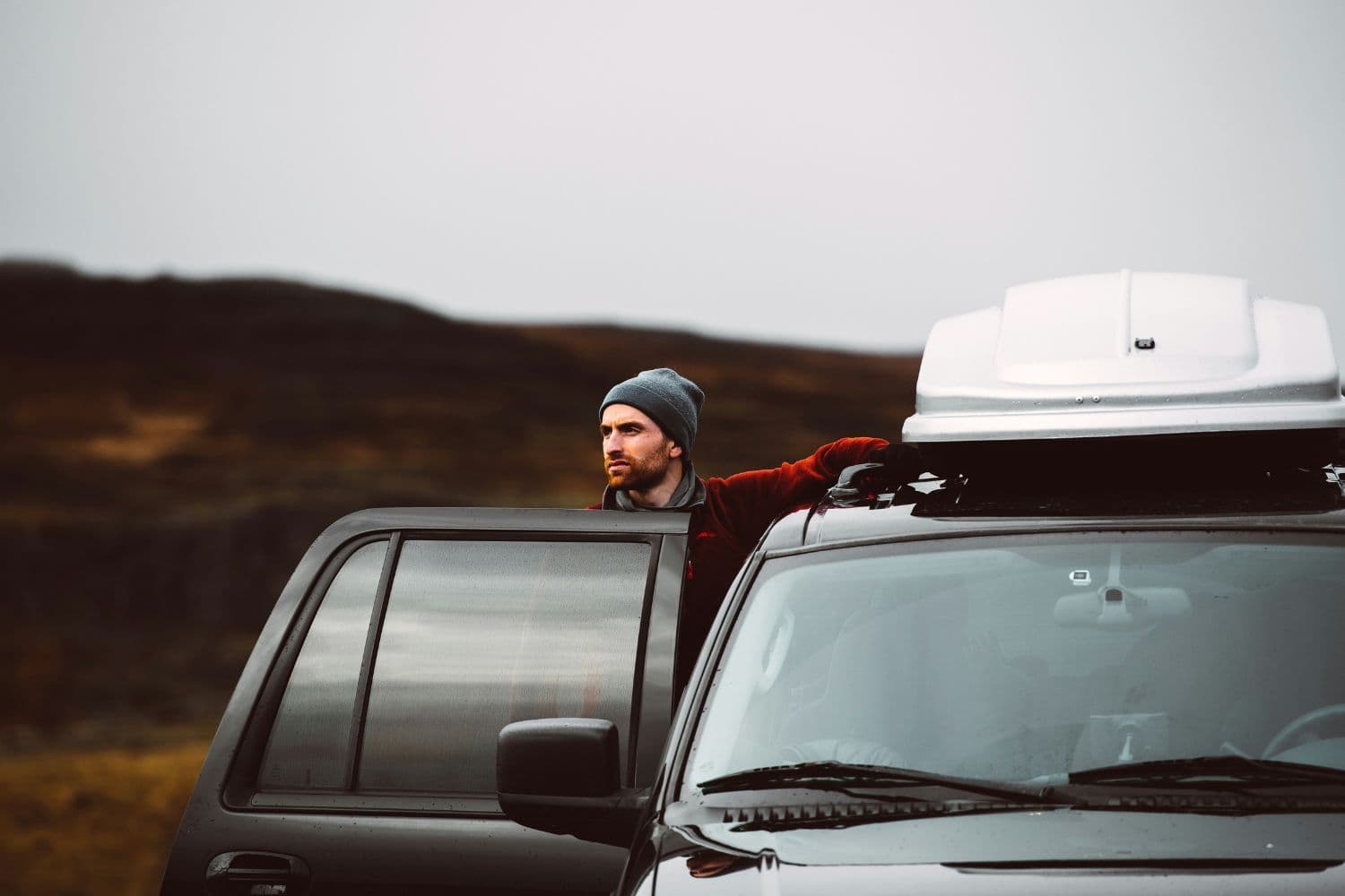 Personne adossée au toit de sa voiture noire dotée d’un coffre de toit blanc, portière droite ouverte, dans un paysage ressemblant à l’Islande