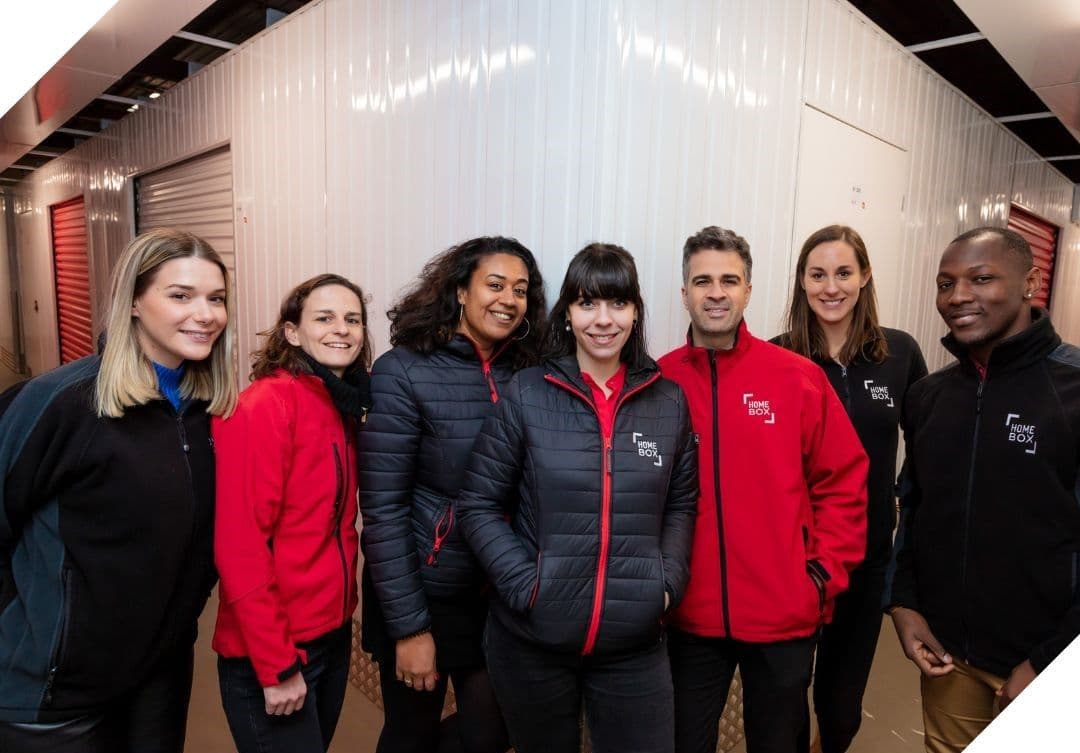 Équipe de conseillers HOMEBOX posant ensemble dans le couloir d’un centre de stockage, portant les couleurs de l’enseigne.