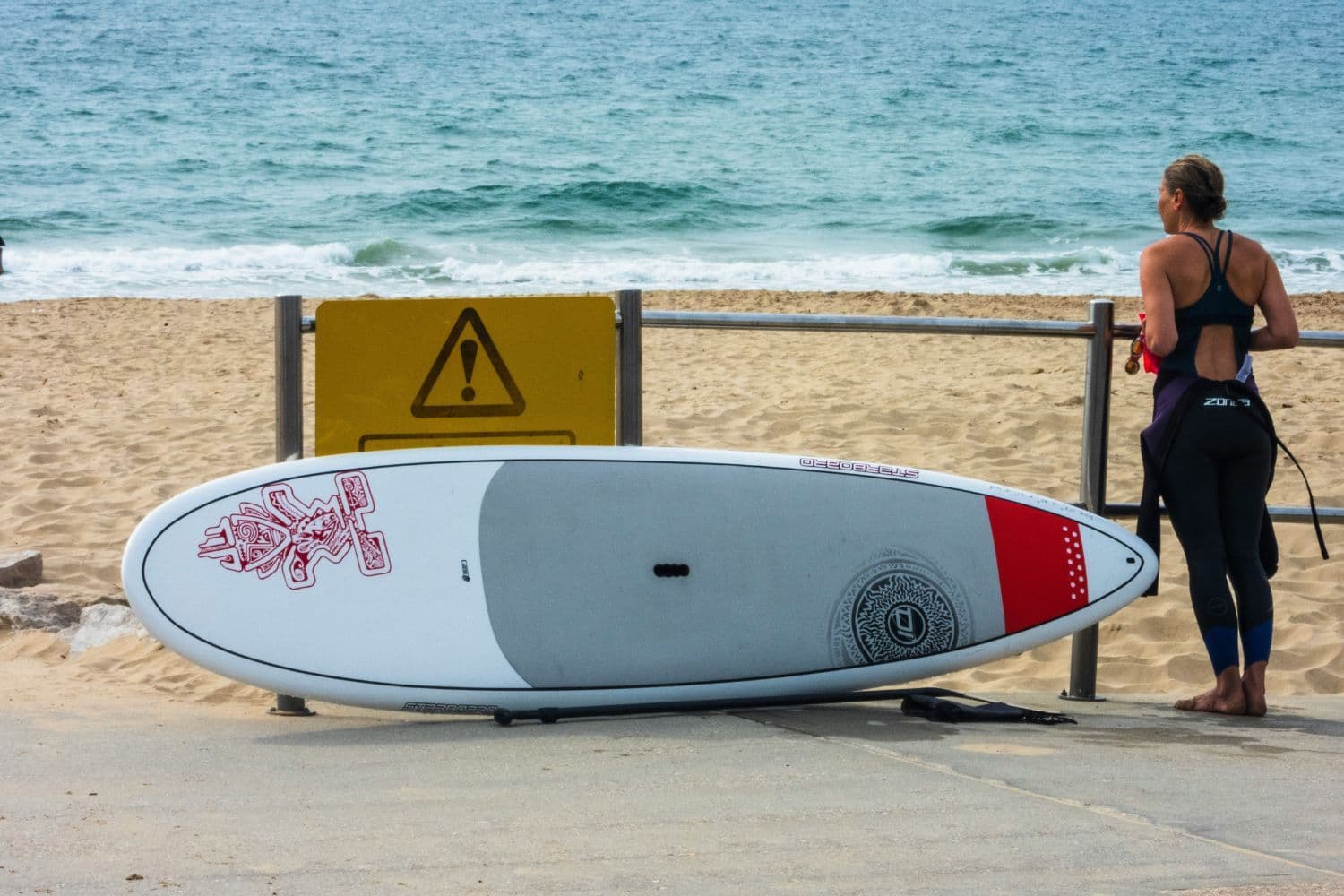 Personne en combinaison sur une plage à côté de sa planche de surf posée au sol à l’horizontal