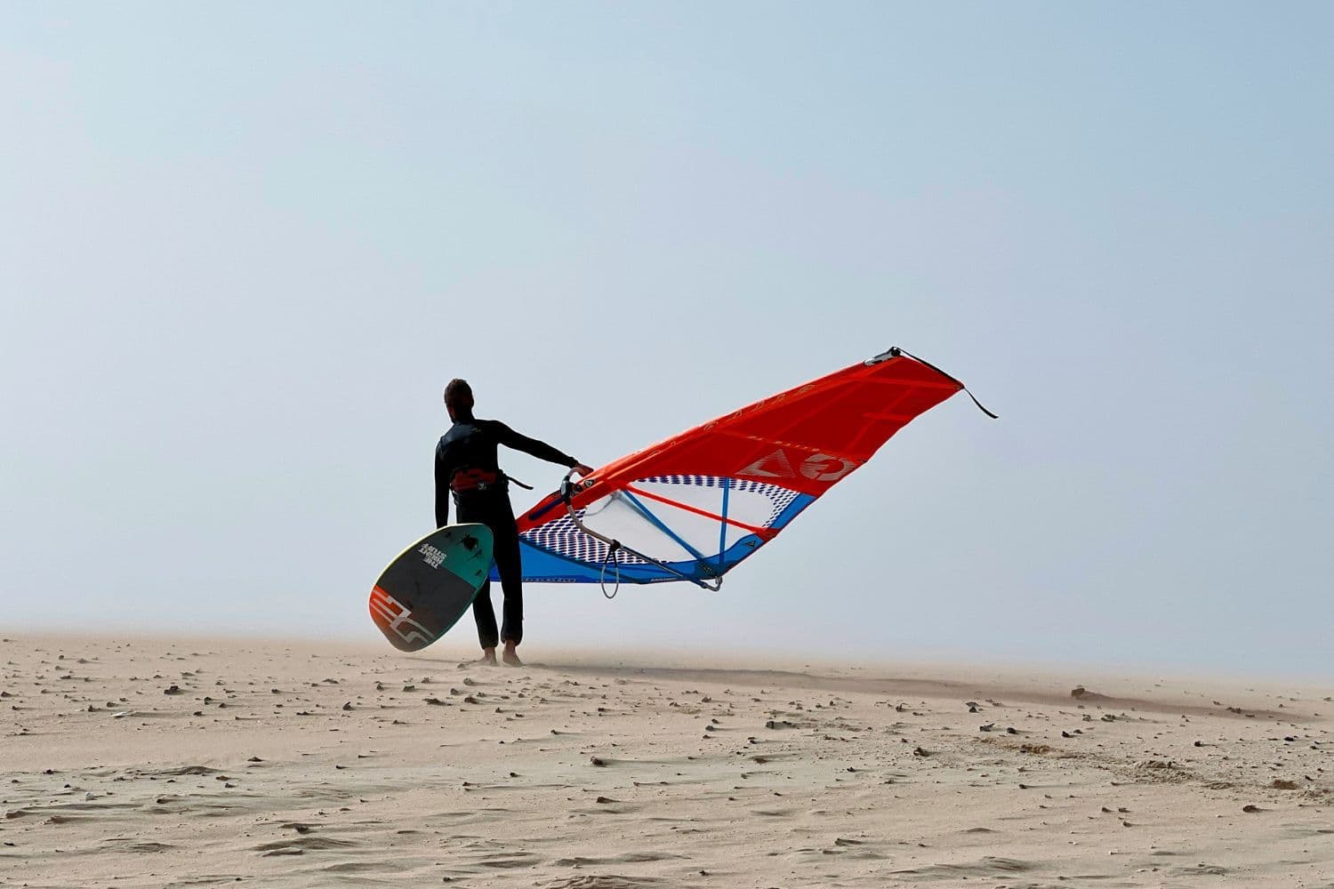 Personne de dos portant une planche à voile sur une étendue de sable