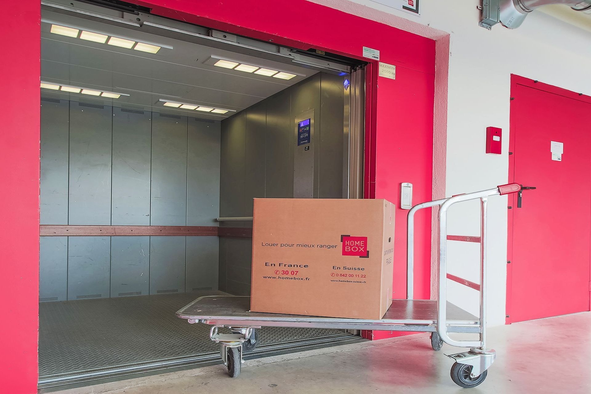Monte-charge ouvert avec chariot de manutention chargé d'un carton dans un couloir rouge du centre HOMEBOX Vannes Nord