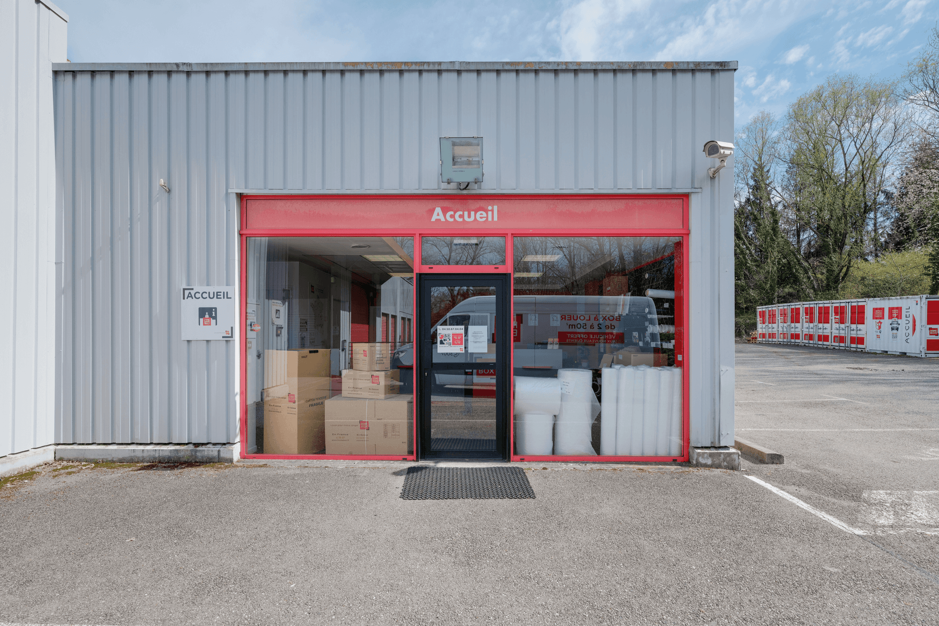 Façade et entrée de l’accueil du centre HOMEBOX Annecy, zone des Vernes à Cran-Gevrier, avec cartons et fournitures visibles à l’intérieur.