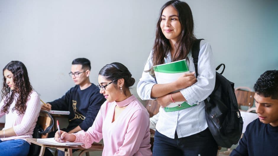 Salle de classe avec élèves assis et concentrés, une élève debout tenant des cahiers