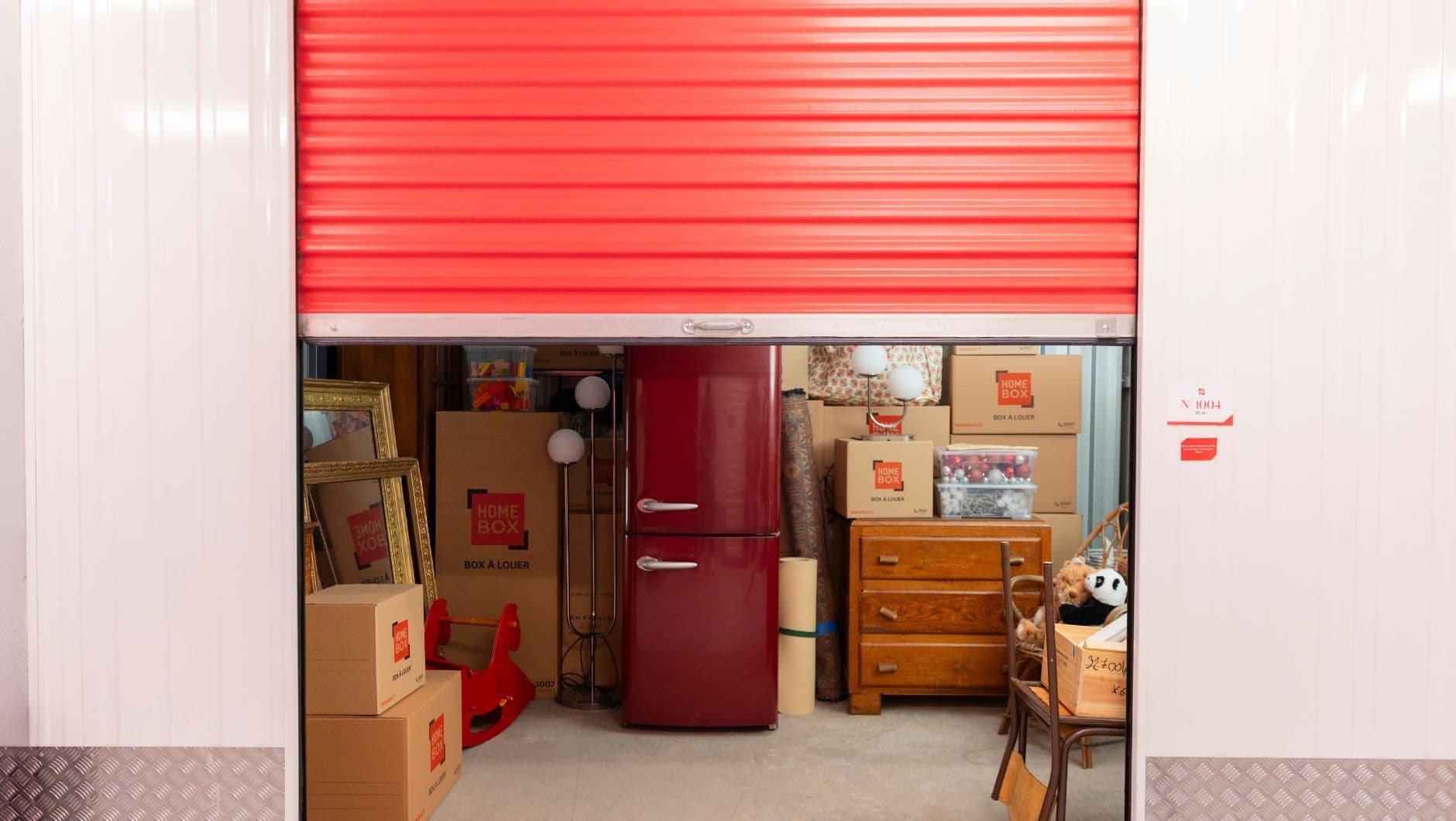 Vue sur un box de stockage de face semi ouvert rempli de cartons HOMEBOX, d’un frigo, d’une commode en bois, de chaises et de miroirs