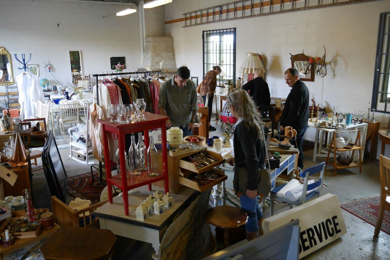 Plusieurs personnes déambulant dans une brocante, nombreux meubles et objets vintage