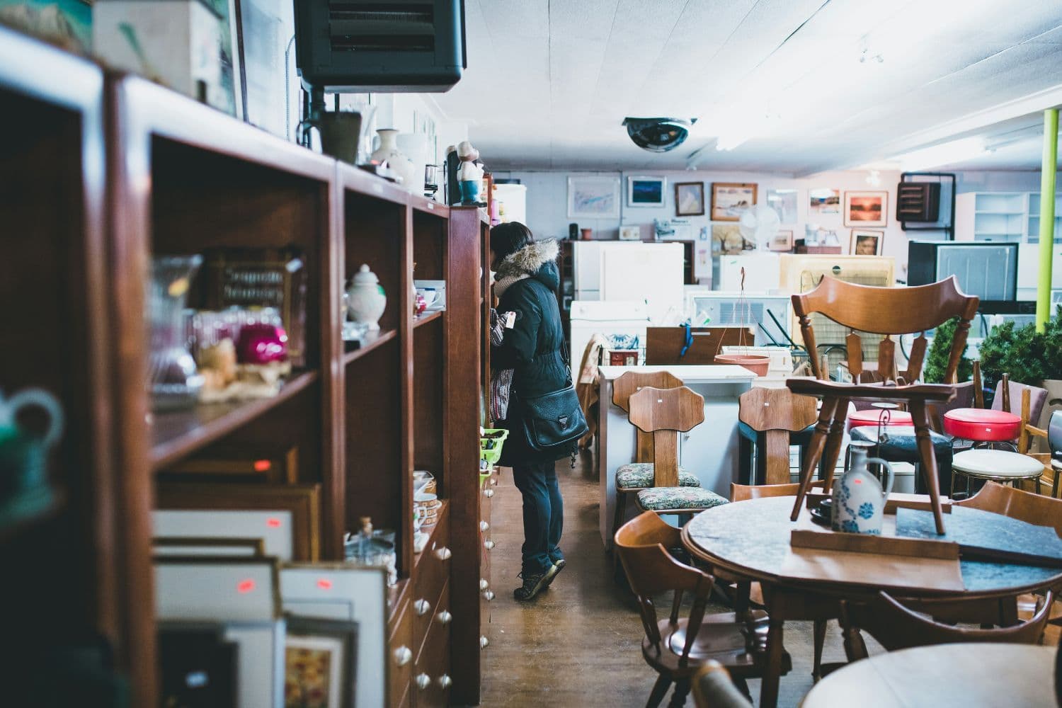 Vue sur un dépôt-vente. Tables, chaises et mobilier en bois à droite, étagères avec de petits objets et des cadres à gauche, personne déambulant dans le fond