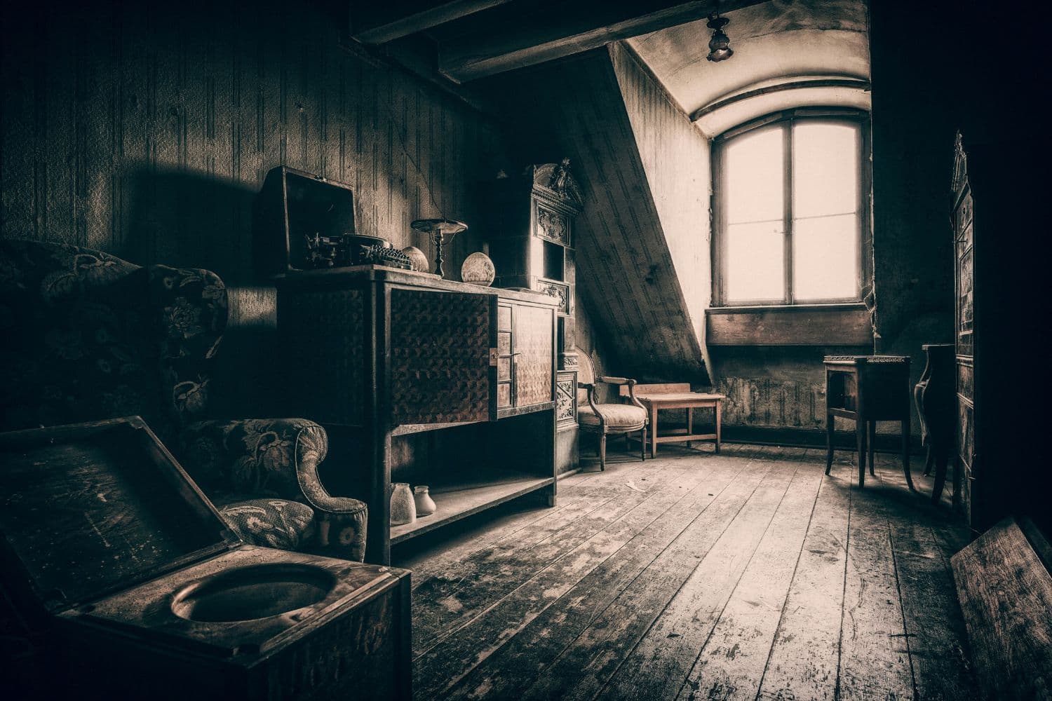 Vue d’un grenier avec, sur le côté, des meubles anciens et, dans le fond, une vaste fenêtre baignée de lumière