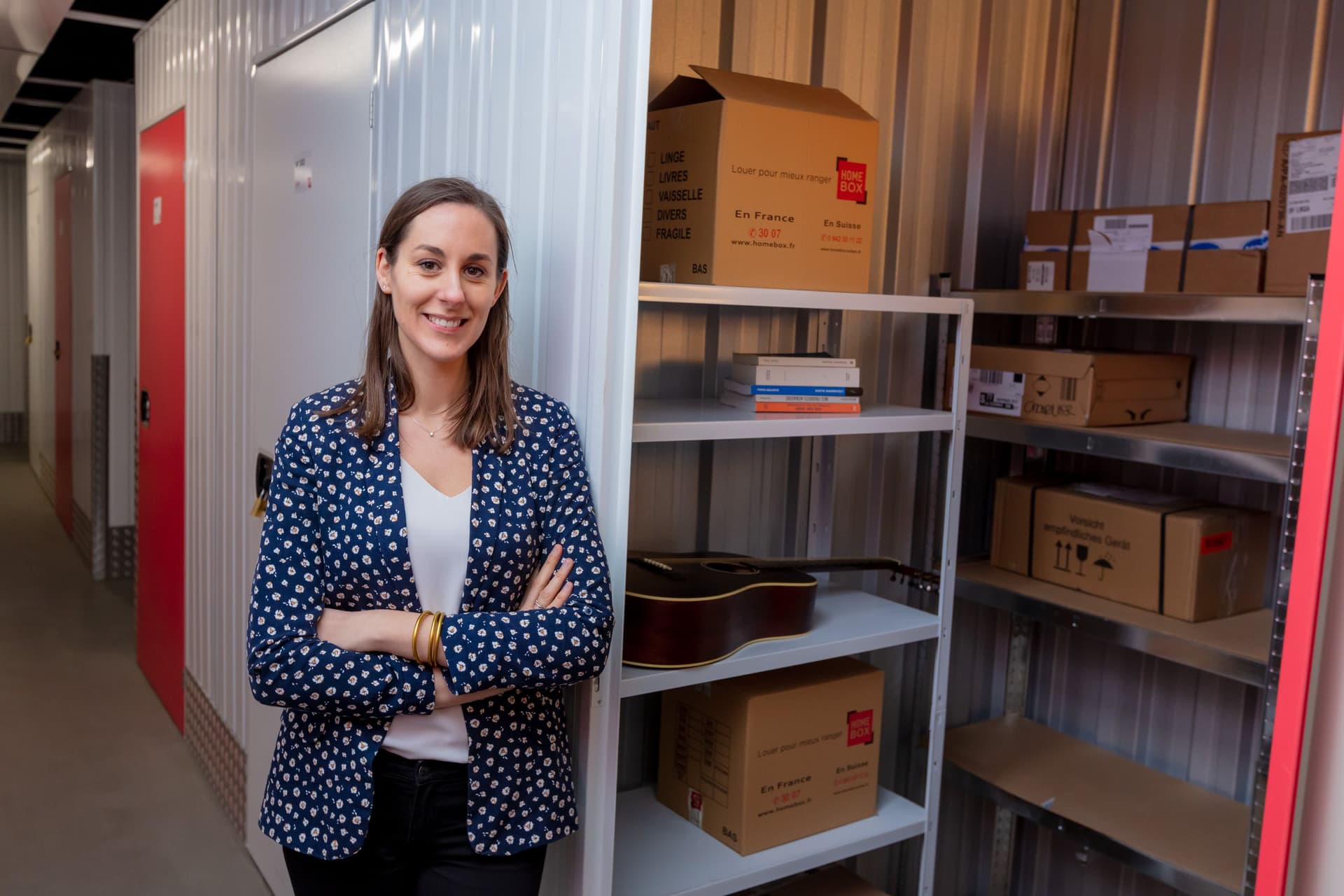 Personne portant une veste bleue à fleurs, appuyée sur le mur de son garde-meuble, cartons HOMEBOX sur des étagères métalliques disposées dans le box