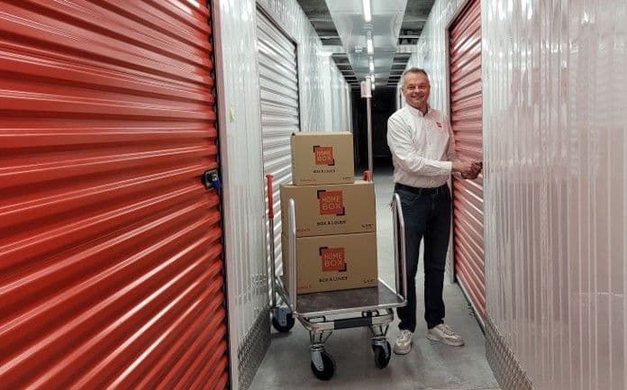 Collaborateur souriant transportant des cartons HOMEBOX sur un chariot dans un couloir de boxes rouges du centre HOMEBOX Nancy